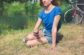 A young woman is sitting by the water in a park and is using a smart phone