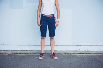 A stylish young woman wearing shorts is standing by a blue wall outside
