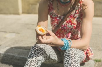 A young woman is sitting on the curb on the street and is eating a scotch egg