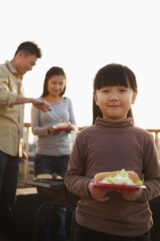 daughter holding plate with hotdog and salad, parents standing next to the barbeque