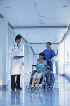 Smiling female nurse pushing and assisting patient in a wheelchair in the hospital, talking to doctor