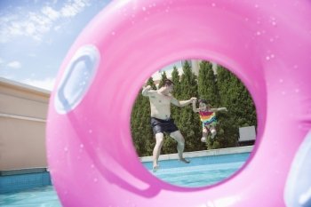 Father and daughter holding hands and jumping into the pool, seen through an inflatable pink tube 