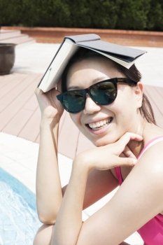 Portrait of smiling woman by the pool in sunglasses holding a book over her head
