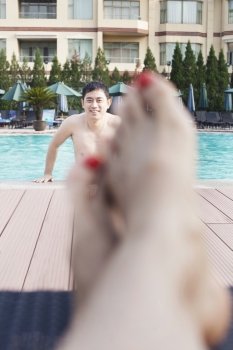 Close up of young woman’s feet by the edge of the pool, man getting out of the water in the background