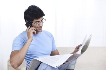 Young man reading newspaper while talking on mobile phone 