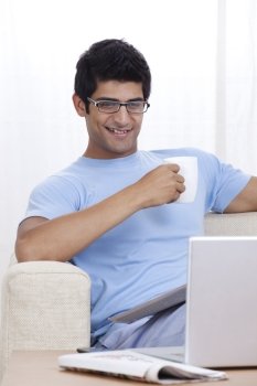Smiling young man having coffee while looking at laptop 