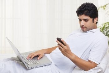 Young man reading text message on cell phone while working on laptop 