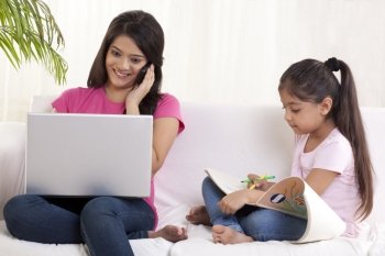 Mother using laptop while talking on phone with daughter sitting besides her and drawing 