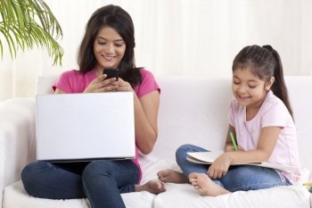 Smiling young woman texting with daughter sitting besides her and drawing 