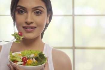 Portrait of happy young woman eating salad of lettuce, cherry tomatoes and mushrooms against glass window 