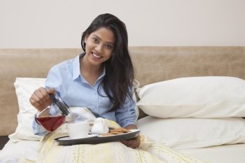 Portrait of beautiful young woman pouring tea in cup while sitting in bed 