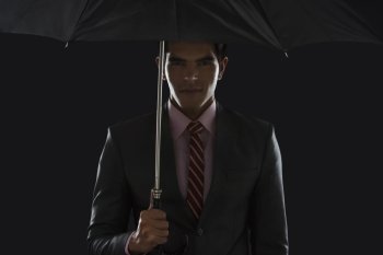 Portrait of confident young businessman holding umbrella against black background