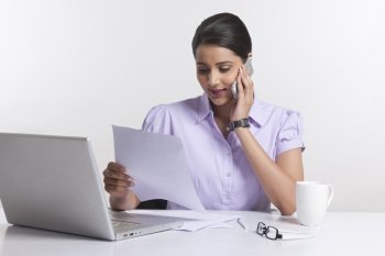 Businesswoman reading document while answering smart phone at desk against white background