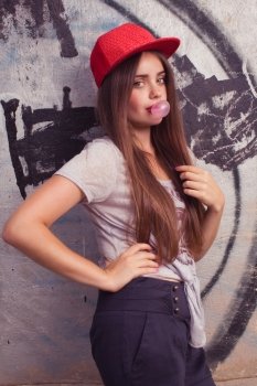 trendy beautiful long haired model posing on graffiti background. Blow bubblegum and show thumb up. red cap. grey t-shirt.