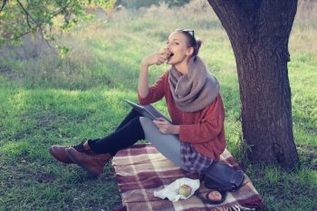 Young trendy woman eating apple and using tablet computer during picnic