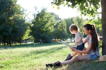 Young stylish mother and toddler reading book at garden during summer fun