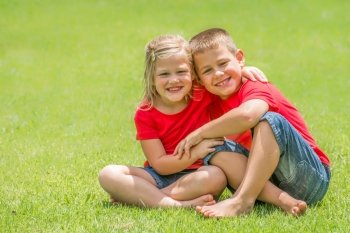 Two children whom are brother and sister are sitting on the green lawn with red t shirts and blue jean shorts with big smiles looking at the viewer at giving each other a big hug, on a beautiful summer day.