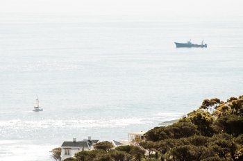 A view of the Atlantic ocean with fishhing trawler and a small private sailing yacht in the water while the tree line and some big private residential houses of Camps Bay are visible in the foreground.