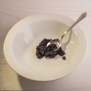 A silver tea spoon lies inside a white ceramic bowl , dipped in to some black caviar, all situated on a white  wooden surface.
