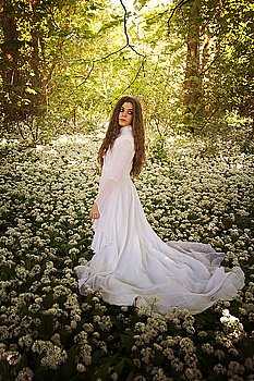 Beautiful woman wearing a long white dress looking over her shoulder whilst standing in a forest of white flowers
