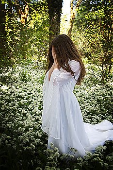 Woman wearing a  long white dress standing in a forest covering her face with her hands