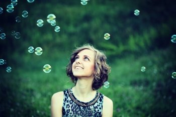 Sweet, happy, smiling blonde girl sitting on a grass in a park playing with bubbles and laughing, having fun. Lifestyle, outdoors