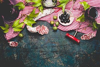 Italian appetizer snack on red cell cotton tablecloths with salami, olives and red wine, dark rustic background, top view, border