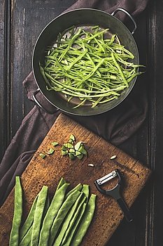 Green french beans cooking. Slices  Green french beans in cooking pot and vegetable peeler on dark rustic wooden background, top view