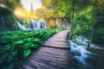 Waterfall and wooden path in green forest in Plitvice Lakes, Croatia at sunset in summer. Colorful landscape with trail in blooming park, trees, water lilies, river, sunbeams in spring. Trail in woods