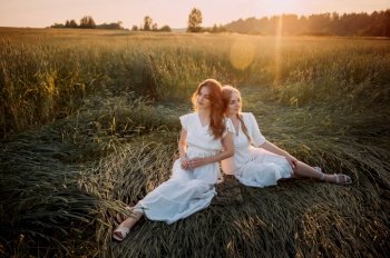 The setting is outdoors, with grass and a clear sky in the background. It exudes a summery vibe with a sense of nature and sunlight.. The photo depicts two women sitting in a field, possibly wearing wedding dresses. 5681