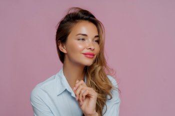 Thoughtful woman in light blue shirt with hand on chin, looking away, against a pink backdrop
