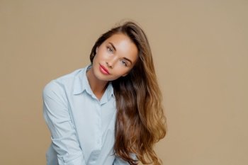 Sultry woman with voluminous hair in blue blouse tilting head on a beige background