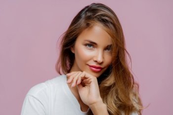 Captivating woman with a hand on her chin, wavy hair, wearing a white top on a pink backdrop