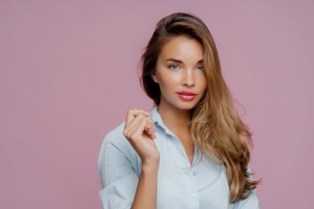 Stylish woman with wavy hair in light blue shirt, touching collar, against a pink background