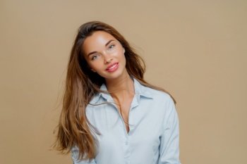Charming woman with tousled hair and open light blue shirt gazes softly on a beige backdrop.