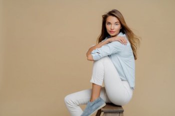 Confident woman in blue shirt and white jeans sitting on stool, looking at camera on a beige background