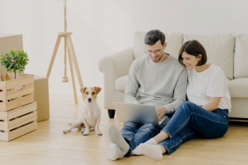 A cheerful couple and their dog in a bright living room with packed moving boxes, bonding over a laptop