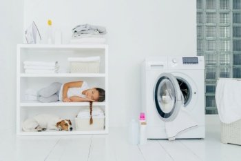 Child and dog napping by laundry machine with ironed clothes and detergents on shelves