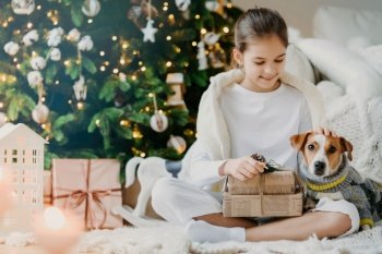 Smiling girl unwrapping a Christmas gift with her dog in a festive sweater, by a beautifully decorated tree