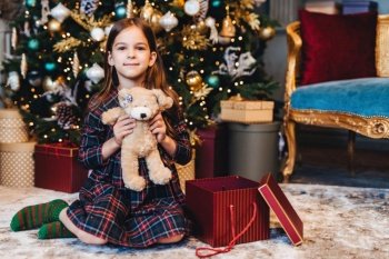 Content girl in plaid pajamas holding a teddy bear, with a Christmas tree and presents in an ornate room