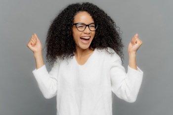 Joyful woman with curly hair and glasses celebrating a victory on a grey backdrop
