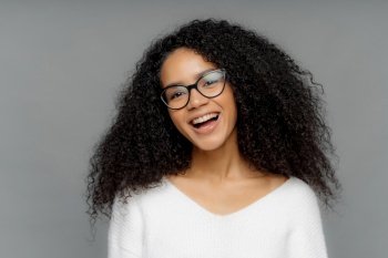 Radiant young woman with glasses and curly hair laughing joyfully on a grey background