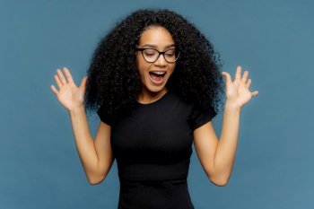 Joyful young woman with glasses and curly hair, excitedly raising hands, on blue background.