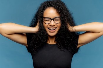 Happy woman with glasses and curly hair, laughing with hands on head, blue background