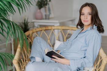 Relaxed woman in striped pajamas reclining in a rattan chair with a book, serene home atmosphere with green plants