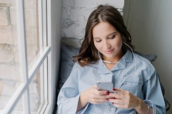 Content young woman in pajamas using smartphone with earphones by the window, enjoying a relaxed moment at home
