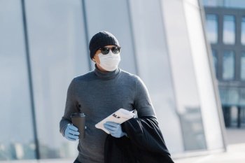 Man with coffee and newspaper walking in the city, wearing a mask and gloves for safety
