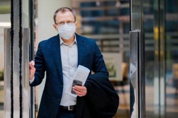 Professional man exiting a building holding a smartphone and newspaper, wearing a mask