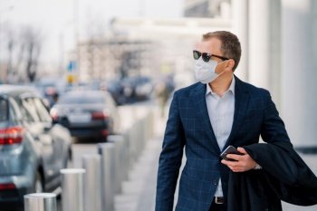Stylish businessman in a suit and mask walking in the city, holding a smartphone