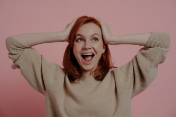 Excited redhead woman holding her head in awe, wearing a cozy sweater, against a pink backdrop.
